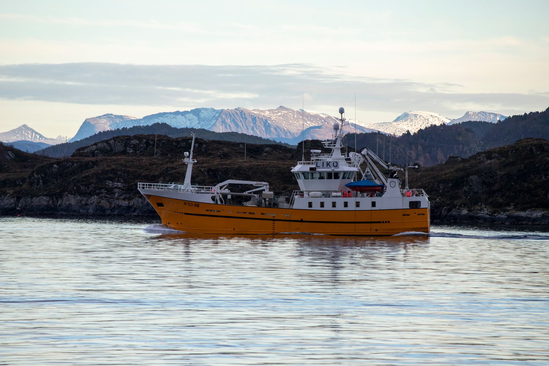 Gul arbeidsbåt på vei gjennom fjord med snødekte fjell bak